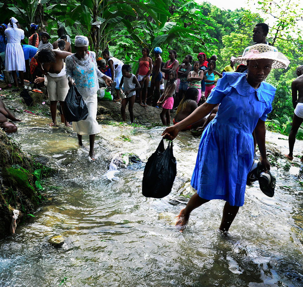 (C)Stanley Greene / NOORIMAGES Ville Bonheur, Saut Deau, Haiti. July 2010. A young girl in blue dress with a white bonnet hat Crossing the cleansing stream at Saut d'Eau